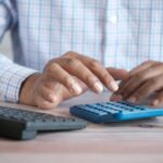 Pessoa usando calculadora sobre uma mesa de trabalho ao lado de uma caneca de café
