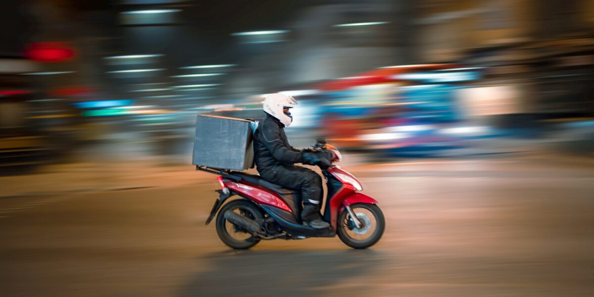 Entregador em scooter vermelha circulando por rua de cidade à noite