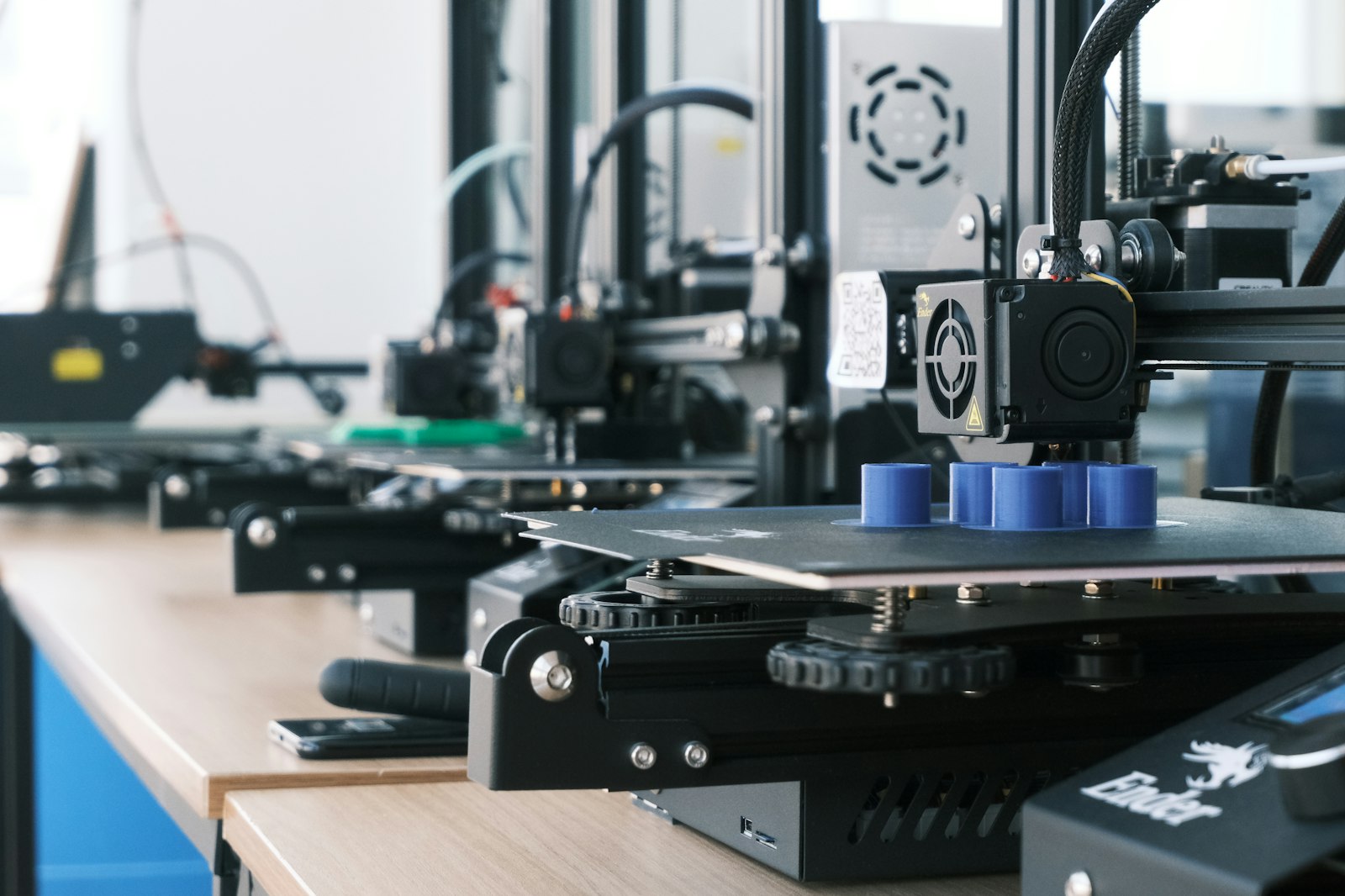 Fileira de impressoras 3D criando peças plásticas azuis sobre uma bancada de madeira